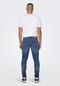 Jeans de denim azul para hombre con un diseño ajustado, que cuentan con bolsillos traseros y un parche de cuero marrón en la cinturilla. Estilizados con una camiseta blanca.