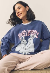 Blauwe sweatshirt met grafische print van drie witte konijnen en de tekst "BUNBELIEVABLE" in roze. Zacht materiaal, ontspannen pasvorm, ronde hals.