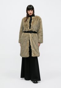 Kunstpelzmantel in Hellbraun mit langen Ärmeln und offenem Vorderteil, ausgestattet mit einem schwarzen Gürtel in der Taille. Wird über einem schwarzen Kleid getragen.