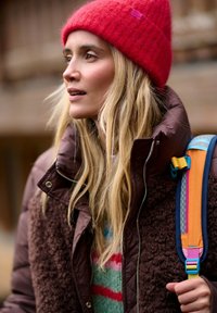 Hnedá nadýchaná bunda s textúrou fleece, červená pletená čiapka a farebný ramenný popruh s oranžovými, modrými a ružovými akcentmi. Pod ňou zelený sveter.