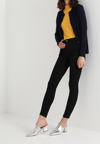 Jeans skinny negros combinados con un suéter de cuello alto acanalado amarillo brillante y un cárdigan azul marino. Mules plateados de punta completa el conjunto.