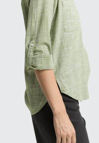 Camicia a quadri verde con maniche arrotolate, dotata di taschino sul petto e orlo posteriore leggermente più lungo per una vestibilità rilassata.