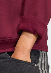 Maroon sweatshirt with a rolled cuff, made from soft fabric. Paired with dark gray jeans featuring striped side detailing and pocket access.