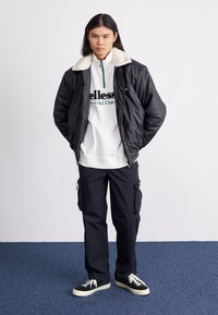 Chaqueta negra acolchada con cuello de piel sintética, pulóver blanco con cremallera y texto verde, pantalones cargo negros y zapatillas negras con suelas blancas.