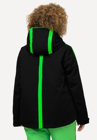 Schwarze isolierte Jacke mit einem grünen vertikalen Streifen in der Mitte und entlang der Kapuze. Verfügt über eine lässige Passform und eine verstellbare Kapuze.