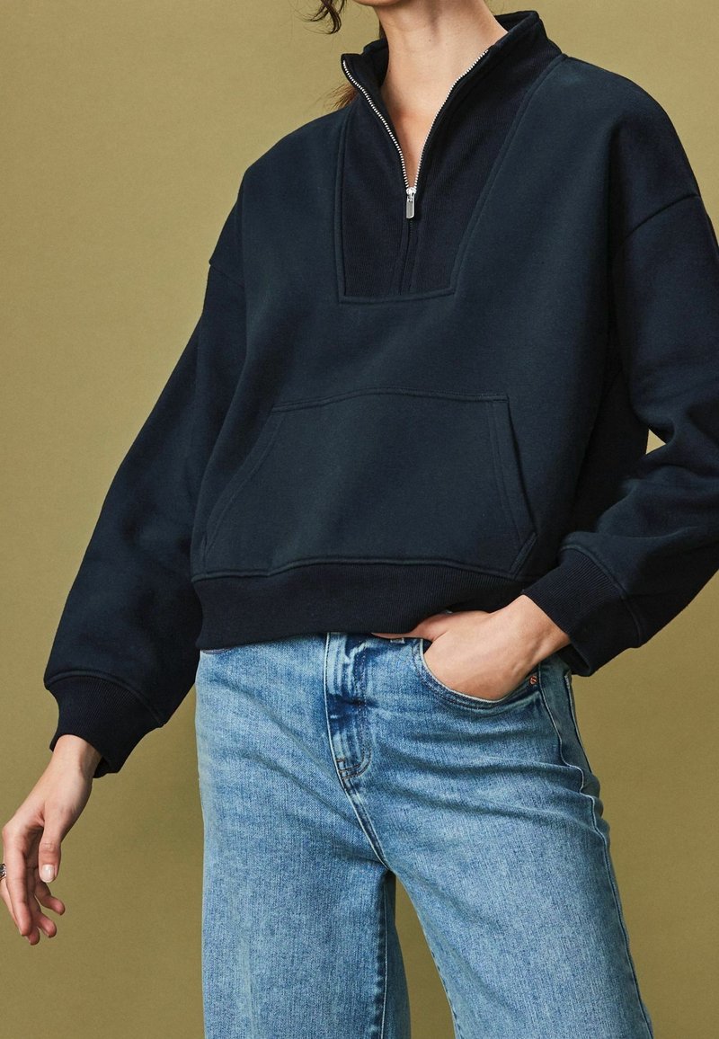 Black fleece pullover with high collar, half-zip front, and large front pocket. Worn with light blue denim jeans against a muted background.