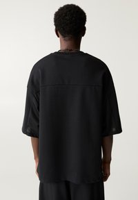 Maglietta nera oversize con maniche corte in rete, scollatura rotonda e vestibilità rilassata, con una texture liscia e dettagli minimi.