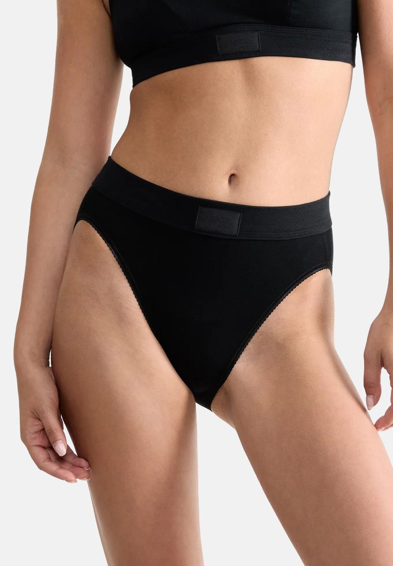 Femme portant un bas de bikini noir taille haute et un crop top noir assorti sur un fond clair uni.