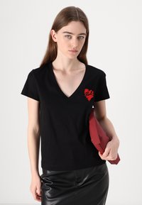 Juodas medvilninis marškinėliai su V formos iškirpte ir raudonu siuvinėtu širdies grafiku ant krūtinės. Modelis laiko raudoną delninę, vilkėdama juodą odinę sijoną.