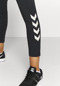 Schwarze Sportleggings mit weißen Chevron-Mustern an der äußeren Oberschenkelseite, kombiniert mit schwarzen Nike-Schuhen, die einen weißen Swoosh haben.