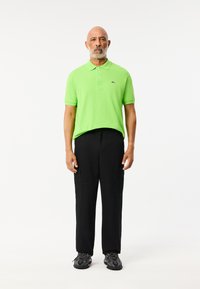 Felgroen polo-shirt met een drieknops sluiting, zwarte cargo-broek met zakken en zwarte sneakers. Effen witte achtergrond.