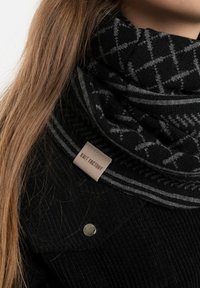 Zwarte gebreide sjaal met een grijs geometrisch patroon, voorzien van een beige label met de tekst "KNIT FACTORY." Materiaal lijkt zacht met een gestructureerd oppervlak.