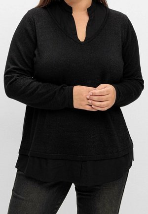 Persoon die zwarte lange mouwen in laagjes draagt met een V-hals over een zwarte broek, staand met de handen samengevouwen ter hoogte van de taille.