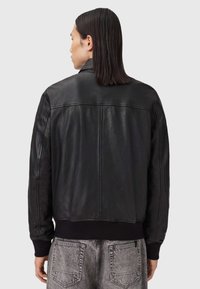 Person med rakt svart hår iklädd en svart läderjacka och grå denimjeans, visad bakifrån mot en enkel bakgrund.