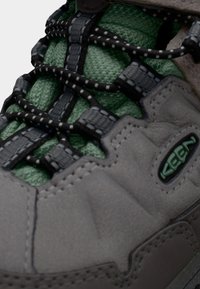 Γκρι πεζοπορικό παπούτσι με υφασμάτινο συνθετικό επάνω μέρος με υφή, επένδυση από πράσινο ύφασμα και μαύρο σύστημα κορδονιών με αντανακλαστικές λεπτομέρειες. Το λογότυπο είναι ορατό στο πλάι.