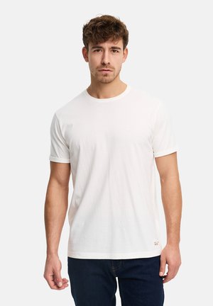 Jonge man met kort, krullend haar, gekleed in een simpele witte korte mouw T-shirt en donkere spijkerbroek, tegen een effen achtergrond.