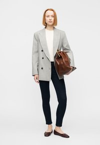 Blazer gris croisé porté sur un haut crème, associé à un jean foncé et un sac en cuir marron. Le modèle, en chaussures plates marron, se tient devant un fond uni.