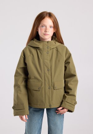 Meisje met lang rood haar, gekleed in een olijfgroene hoodie en blauwe jeans, staand tegen een lichtgrijze achtergrond.