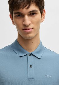 Ljusblå polo skjorta i strukturerad tyg, med klassisk krage, två svarta knappar och broderad "BOSS" logotyp på bröstet.