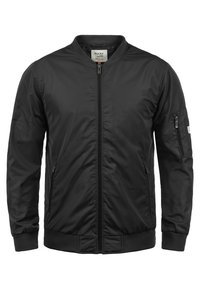 Musta bomber-takki kiiltävästä kankaasta, jossa on etuvetoketju, ribboden hihansuut ja vetoketjullinen tasku vasemmassa hihassa.