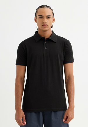 Joven con trenzas cornrows que lleva una camisa polo negra de manga corta y pantalones oscuros, de pie frente a un fondo blanco liso.