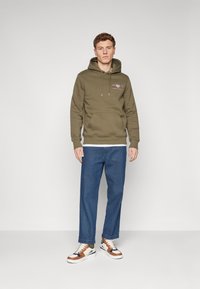 Felpa con cappuccio verde oliva, dotata di tasca frontale, abbinata a jeans in denim blu a vita alta e scarpe da ginnastica multicolori.