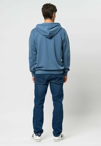 Blauwe hoodie met een gladde textuur, gecombineerd met donkere spijkerbroeken. Sneakers hebben een witte basis en gekleurde strepen aan de zijkant.
