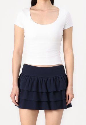 Kobieta ubrana w dopasowaną białą bluzkę z krótkimi rękawami i okrągłym dekoltem oraz granatową mini spódniczkę z falbankami w warstwach, stojąca na jednolitym tle.