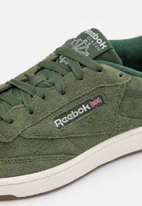 Zelený semišový teniska s výraznými tkaničkami, strukturovaným povrchem, bílými detaily loga a kontrastní gumovou podrážkou.