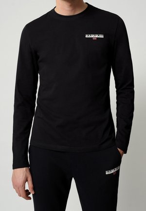 Mies, joka pukeutunut mustaan pitkähihainen paitaan ja housuihin, joissa on pieniä valkoisia Napapijri-logoja rinnassa ja reiden yläosassa, seisoo yhdellä kädellä taskussa.