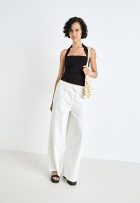 Black halter top paired with loose, white, high-waisted linen pants. Model carries a crocheted bag with floral accents. Black heeled sandals.