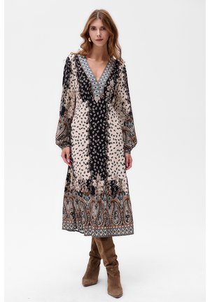 Kobieta z falującymi włosami, ubrana w wzorzystą sukienkę midi z długim rękawem i dekoltem w szpic, oraz brązowe zamszowe kozaki do kolan, stojąca na tle białym.