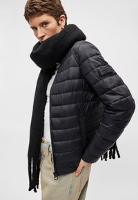 Juodas paminkštintas švarkas su užtrauktuku, su tekstūruotu juodu šaliku ir logotipo šašlu ant rankovės. Dėvimas ant baltos marškinės ir šviesių džinsų.