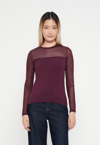 Långärmad topp med en åtsittande vinröd kropp och genomskinliga meshaxlar. Slät tyg med rund hals och kontrasterande texturer. Matchas med mörka jeans.