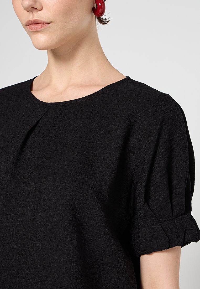 Schwarze Bluse mit rundem Halsausschnitt, kurzen puffärmeligen Ärmeln mit aufgerollten Bündchen und einem strukturierten Stoff.