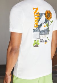 Camiseta blanca con un girasol, planta pequeña, zapatos Nike y el texto "Nike" y "Grow Your Sole" en la parte trasera, usada por una persona con pantalones cortos verdes.