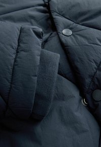 Jachetă pufosă de culoare bleumarin, cu o textură netedă, dotată cu închidere cu capse și manșete striate. Accentele includ un detaliu cu inel metalic.