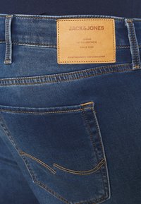 Zadní kapsa a kožená nášivka značky na modrých džínech s oranžovým prošíváním, zobrazující "JACK & JONES" a detaily sloganů.