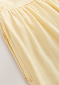 Tessuto di cotone giallo pallido con una consistenza liscia, caratterizzato da dettagli arricciati lungo la vita e una vestibilità leggermente rilassata.