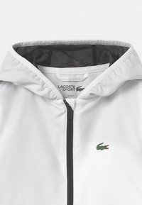 White windbreaker jacket with a black zipper, mesh-lined hood, and a small green crocodile logo on the chest. Textured fabric.