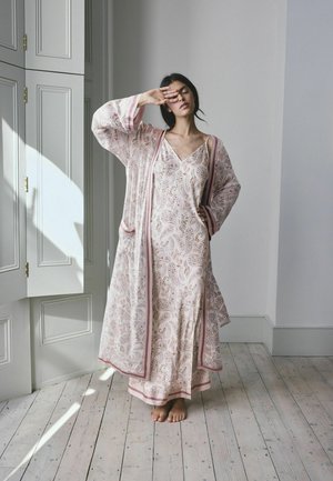 Femme debout pieds nus sur un sol en bois portant une longue chemise de nuit fleurie assortie et un peignoir avec bordure rose, se couvrant les yeux d'une main.