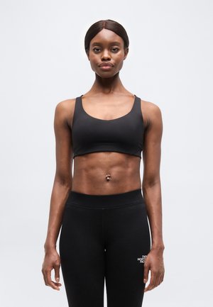 Γυναίκα σε φόρμα, ντυμένη με μαύρο αθλητικό σουτιέν και κολάν, στέκεται με το πρόσωπο προς τα εμπρός μπροστά σε απλό λευκό φόντο.