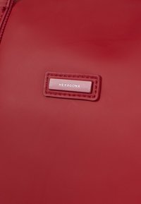 Rood synthetisch materiaal met een gladde textuur, voorzien van een rechthoekig label met het merk "HEXAGONA" erop geperst. Ritsdetail zichtbaar.