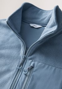 Jasnoniebieska kurtka polarowa z podniesionym kołnierzem, głównym zamkiem częściowo rozpiętym oraz dwoma zapinanymi na zamek kieszeniami na piersi z pętlami do uchwytu, z widoczną metką marki wewnątrz.