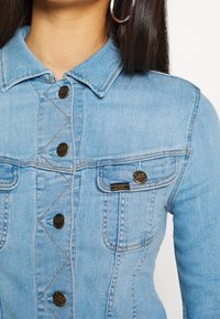 Light blue denim jacket with metal buttons and pocket flap, worn by a person with long dark hair and large silver hoop earring.