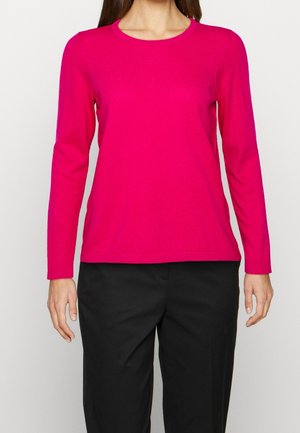 Femme portant une chemise à manches longues rose vif et un pantalon noir, debout devant un fond blanc uni.