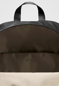 Fekete hátizsák sima szövet külsejével és bézs belsejével. Cipzáras záródással rendelkezik, és egy "lululemon" logó címke található belül.