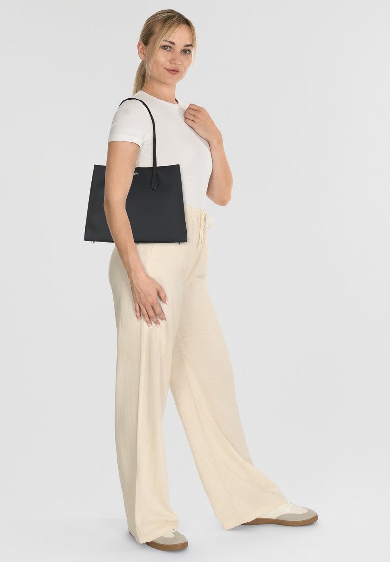 Sac fourre-tout noir au design structuré, texture lisse, avec des poignées épaule, et une esthétique minimaliste, associé à un pantalon beige à jambes larges.