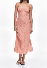Mujer vistiendo un vestido midi floral rosa con tirantes finos y sandalias blancas, de pie contra un fondo blanco.