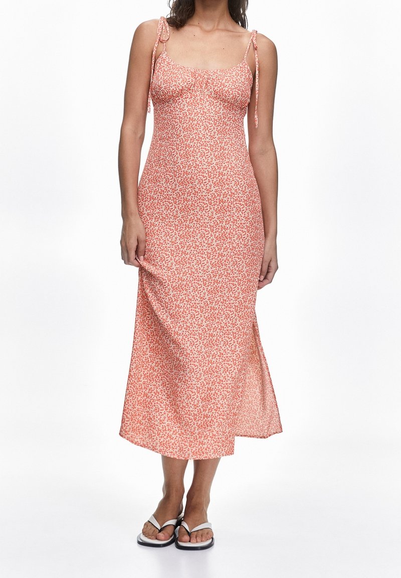 Mujer vistiendo un vestido midi floral rosa con tirantes finos y sandalias blancas, de pie contra un fondo blanco.